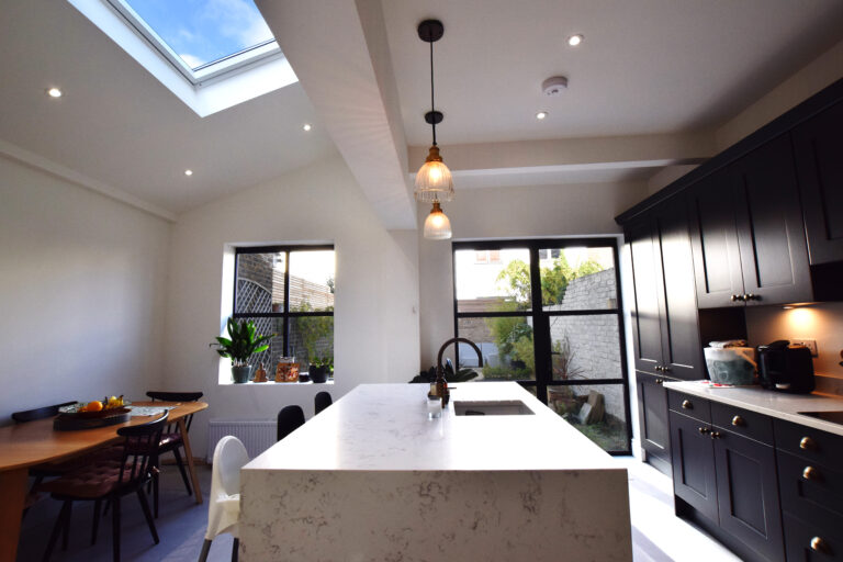 Internal view of a kitchen , the side part has been extended and includes a skylight and the rear elevation has a window and a french door that opens to the rear garden. Plenty of natural ligth highlights the central white marble island. The dark cupboards provides a nice contrast to the area.